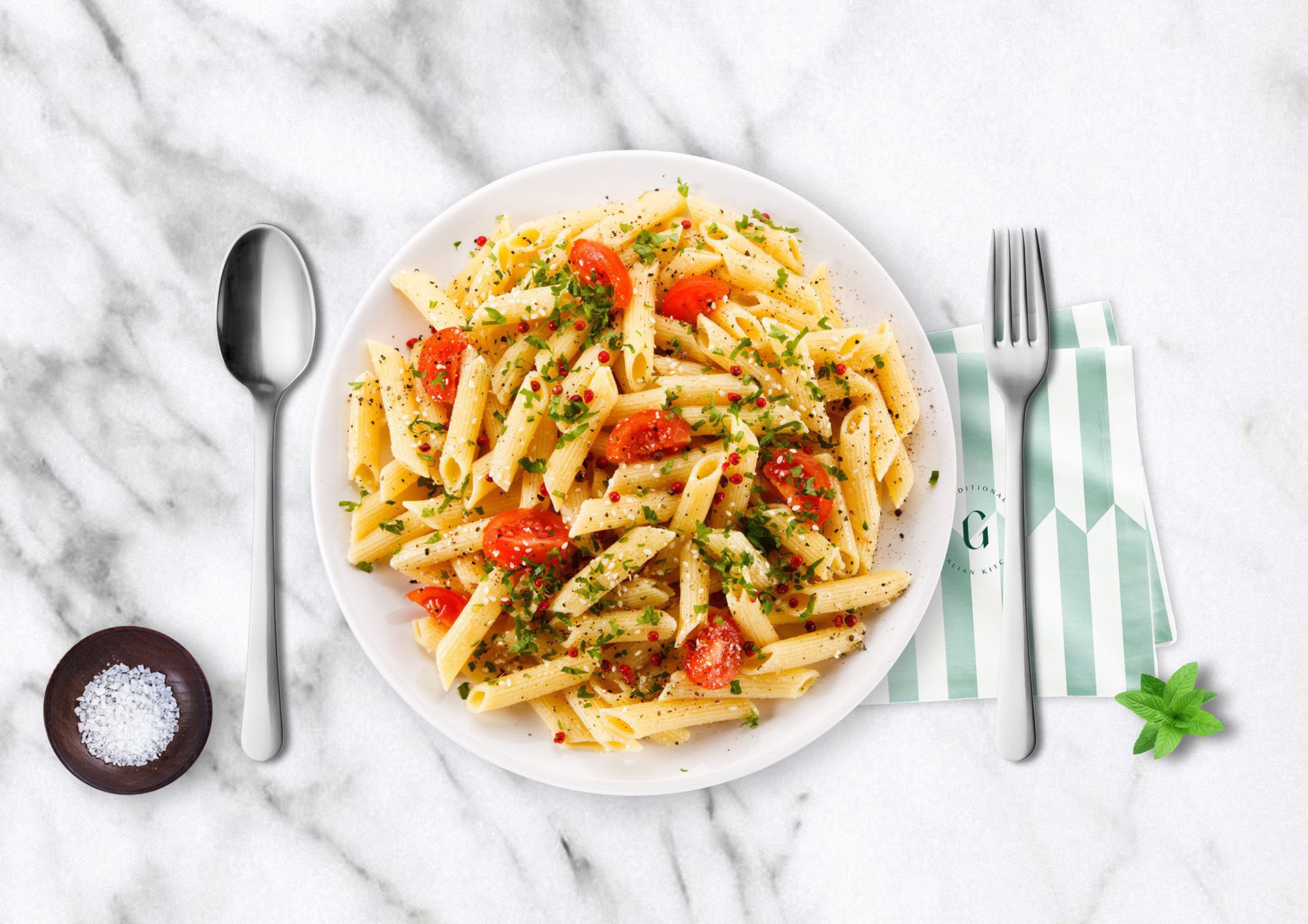 Penne with tomatoes on marble beside a salt bowl and a striped Gaia napkin